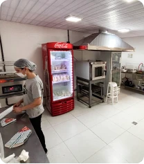 Imagem de uma cozinha de restaurante com equipamentos como geladeira vermelha, forno de micro-ondas e área de preparo, com uma pessoa preparando alimentos.
