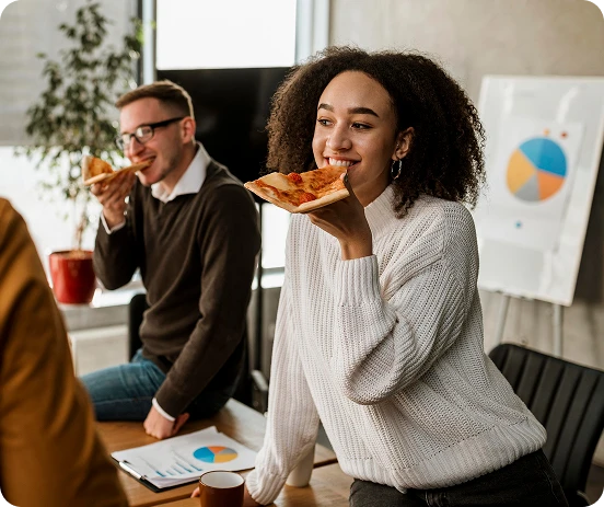 Mulher sorridente segurando uma fatia de pizza em uma reunião de trabalho, com colegas ao fundo e gráficos na parede.
