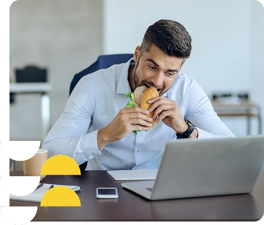 Homem de camiseta social feliz comendo hambúrguer enquanto trabalha no computador, promovendo alimentação saudável na rotina do escritório.