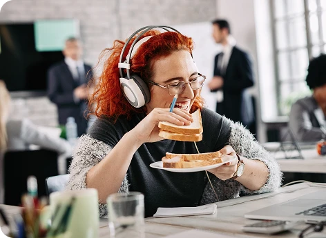 Mulher usando headphones e comendo sanduíche em escritório moderno, focada no trabalho enquanto desfruta de uma pausa, ambiente de escritório com colegas ao fundo.