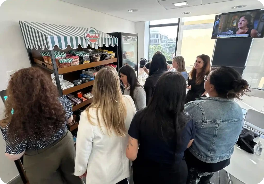 Grupo de mulheres em um evento na loja de snacks, interagindo e conversando em um ambiente moderno com televisão e janela grande ao fundo.