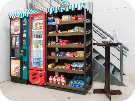 Prateleira de produtos de conveniência com snacks e bebidas ao lado de máquinas de venda automática, localizada junto às escadas em um ambiente interno.